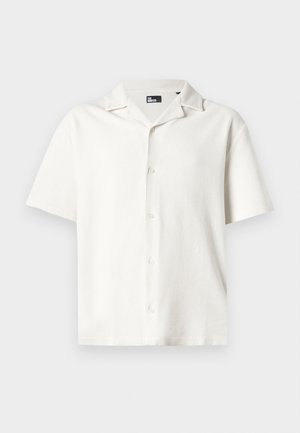 Majica kratkih rukavov v beli barvi, izdelana iz teksturirane tkanine, s kamp ovratnikom in petimi gumbi, ki se ponaša z ohlapnim krojem ter subtilnim vzorcem.