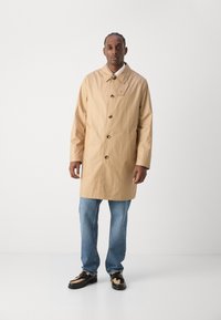 Beige waterdichte trenchcoat met een knoopsluiting, lange mouwen en een puntige kraag. Gepaard met lichtblauwe jeans en zwarte schoenen.