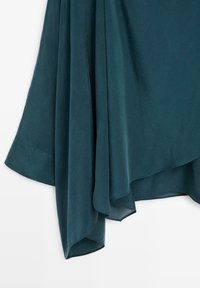 Saltet stof i teal med en blød, glat tekstur, der har et draperet design og en asymmetrisk kant, som skaber flydende linjer og et lagdelt udseende.
