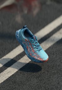 Chaussure de course turquoise avec tige en mesh, accents orange, design léger, semelle courbée et surface texturée, présentée sur un fond de pavé.