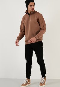 Sudadera de forro polar marrón con cremallera y cuello alto, combinada con pantalones negros ajustados y zapatillas altas en color beige. Modelo con un gorro negro.