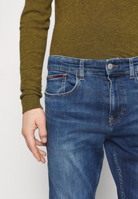 Siniset farkut, joissa on lievä haalistuminen ja punasininen korostus etutaskussa, yhdistettynä vihreään pitkähihaiseen villapaitaan.