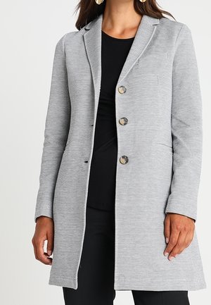 Femme portant un long manteau gris avec trois boutons sur un haut noir et un pantalon noir, visible des épaules aux cuisses.
