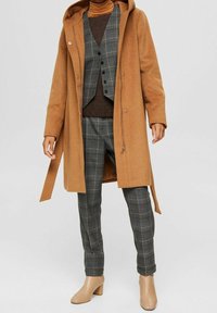 Camelkleurige lange jas over een bruine coltrui, grijze geruite vest en broek, gecombineerd met beige enkellaarsjes op een staande persoon.