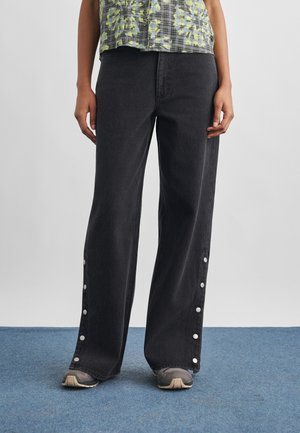 Osoba nosząca czarne spodnie z szerokimi nogawkami z zatrzaskami wzdłuż zewnętrznych szwów, w połączeniu z szarymi sneakersami i wzorzystą bluzką.