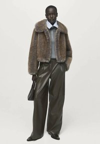 Modelo vistiendo una chaqueta de piel sintética marrón, una capa interior con cremallera en gris claro, pantalones de cuero oscuros de pierna ancha, zapatos negros, sosteniendo un pequeño bolso negro.