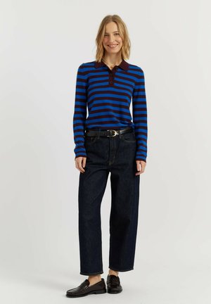 Vrouw glimlachend, draagt een blauw en donkerbruin gestreepte polo met lange mouwen, donkere hoog getailleerde jeans, zwarte riem en zwarte loafers.