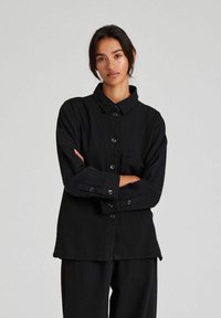 Cămașă neagră cu nasturi, cu guler, mâneci lungi și un buzunar frontal. Are o croială lejeră, cu o textură subtilă și material mat.