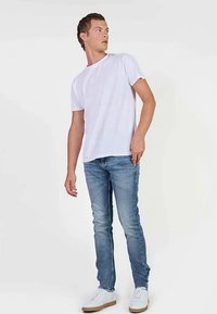 Witte katoenen T-shirt gecombineerd met lichtblauwe spijkerbroek en witte sneakers. Model staat zijwaarts en kijkt over de schouder.