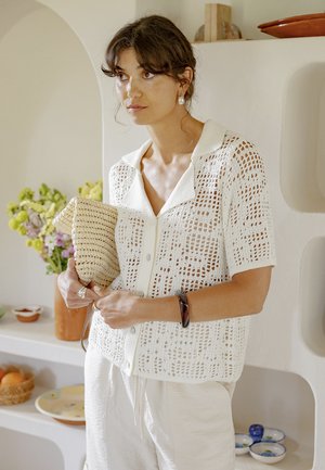 Žena v ľahkej háčkovanej blúzke s krátkym rukávom a bielych nohaviciach drží pletenú kabelku, stojí v interiéri pri poličkách s kvetmi a miskami.