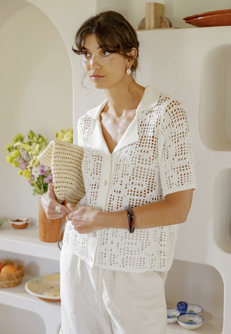 Femme en blouse légère en crochet à manches courtes et pantalon blanc tenant une pochette tissée, debout à l'intérieur près d'étagères avec des fleurs et des bols.