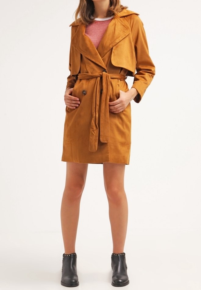 Trench-coat en daim marron avec ceinture, boutons double boutonnage et large col. Porté sur un haut rose et associé à des bottines noires.