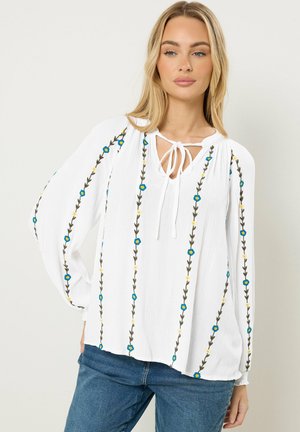 Frau trägt eine weiße Bluse mit vertikaler Blumenstickerei und einem Bindeband am Ausschnitt, kombiniert mit blauen Jeans, steht vor einem schlichten Hintergrund.