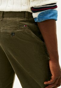 Primo piano di una persona che indossa pantaloni in velluto a coste verde oliva con un piccolo logo rosso, bianco e blu vicino alla tasca posteriore e un maglione crema con maniche in denim blu arrotolate.