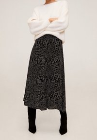 Persoon in een wit donzig sweater, zwarte midi-rok met een wit onregelmatig stippenpatroon, en zwarte haklaarzen, staande met de armen gekruist.