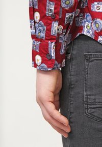 Red shirt with a camera print, featuring a buttoned cuff and smooth fabric. Paired with dark gray denim jeans.