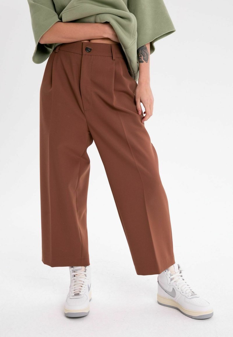 Bruna vida byxor med veck och knappstängning, i kombination med vita och grå höga sneakers. Outfiten kompletteras med en olivgrön topp.