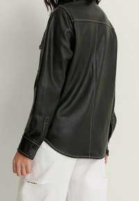 Svart läder skjortjacka med vita sömnadsdetaljer, knappärmar och en rundad fåll. Baksidan har en mjuk yta och avslappnad passform.