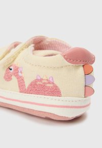 Καμβά παπούτσι σε κρεμ απόχρωση με ροζ κέντημα δεινοσαύρου, υφασμάτινες λεπτομέρειες και πολύχρωμες λαστιχένιες προεξοχές στη φτέρνα. Ροζ ριγωτή εσωτερική επένδυση.