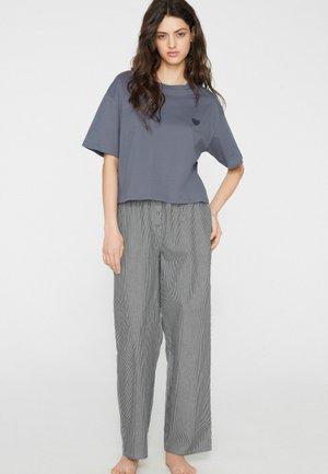 Młoda kobieta o długich falowanych włosach, ubrana w luźny szary krótki t-shirt z małym sercem oraz luźne szare spodnie w prążki, stojąca boso.