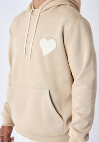 Sudadera beige con cordones, bolsillo frontal y detalle de corazón bordado. Textura suave y corte relajado, adecuada para un uso casual.