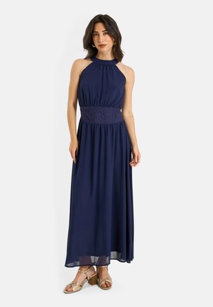 Femme portant une robe longue bleu marine sans manches avec un détail en dentelle à la taille et des sandales à talons blocs ouvertes dorées, debout devant un fond blanc.