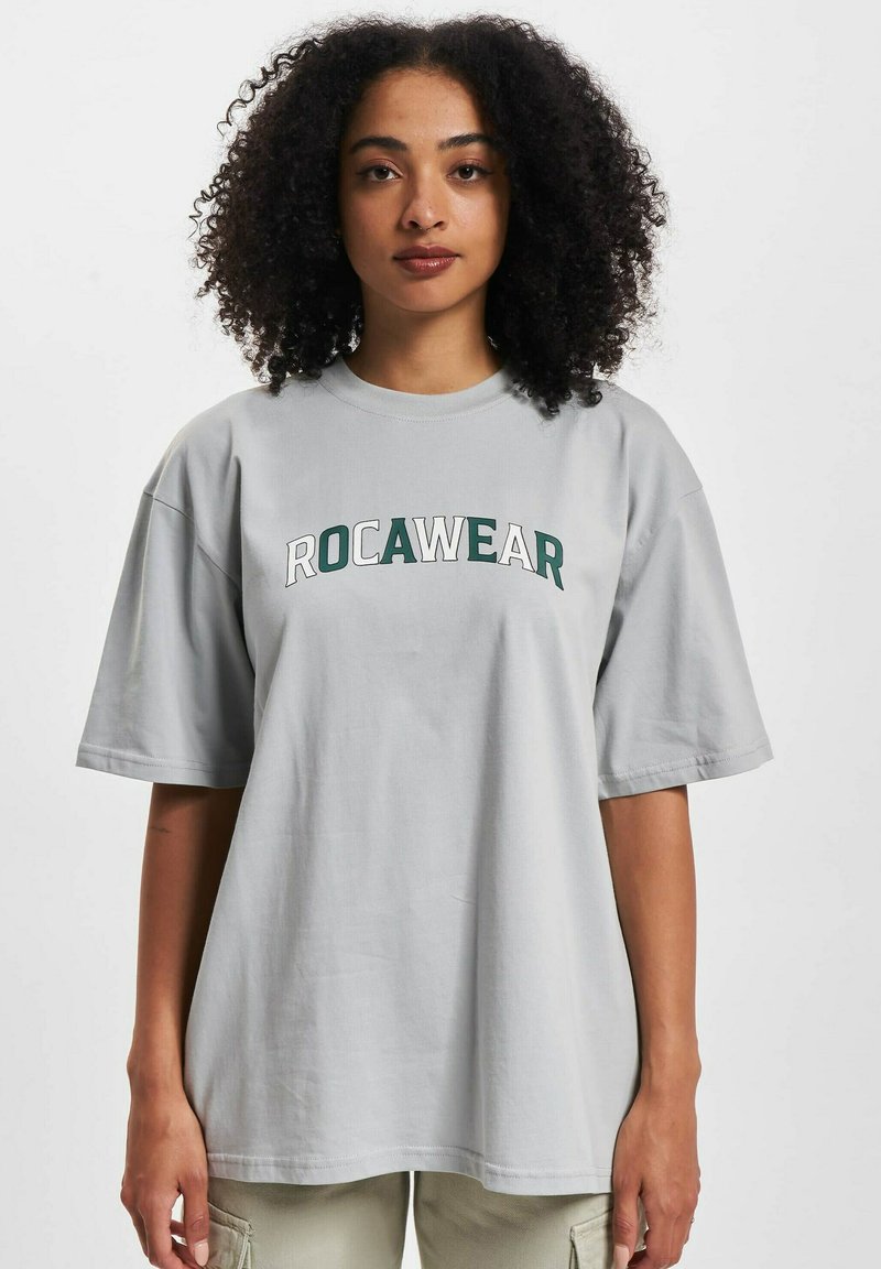 Lys grå oversized T-skjorte laget av bomull, med "ROCAWEAR" trykt i grønt, rund hals og korte ermer.