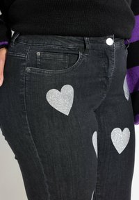 Jeans de mezclilla negra con diseños de corazones de purpurina plateada. Incluyen bolsillos frontales y un cierre de botón plateado.