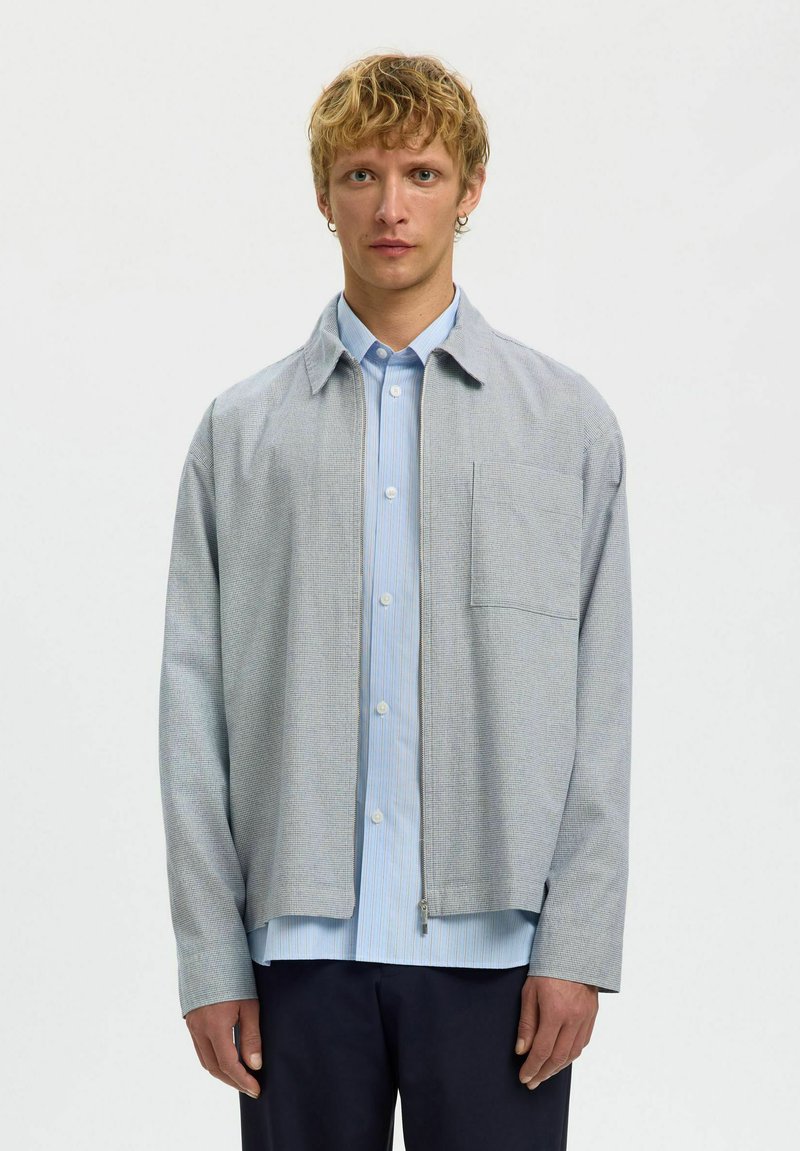 Jeune homme aux cheveux blonds courts et bouclés portant une veste grise claire zippée par-dessus une chemise à boutons bleu clair et un pantalon foncé, faisant face à l'avant.