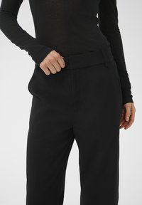 Schwarze, maßgeschneiderte Hose mit weitem Bein, aus glattem Stoff, mit umgeschlagenem Bund und seitlichen Taschen. Das Modell hält den Bund.