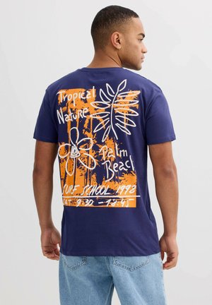 Hombre vistiendo una camiseta azul marino con un gráfico y texto de temática tropical en naranja y blanco, combinado con pantalones vaqueros azul claro, de espaldas.