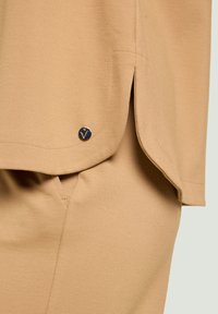 Top beige con una textura suave, una abertura lateral y un pequeño detalle metálico de logotipo cerca del dobladillo. El material parece ser suave y ligero.