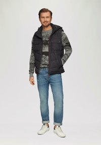 Gilet matelassé noir avec capuche, pull en camouflage gris, jean bleu clair et baskets blanches. Comprend des poches poitrine boutonnées.