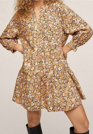 Kvinde iført en løs, blomsterprintet kjole i gult, pink og hvidt med lange ærmer og sorte knæhøje støvler, hænderne på hoften.