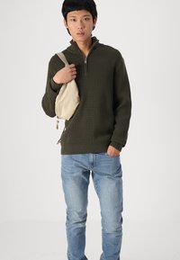 Sötétzöld, textúrált kötött pulóver, cipzáros gallérral, világoskék farmernadrággal és bézs crossbody táskával párosítva. Egyszerű, hétköznapi ruházati dizájn.