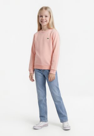 Fille souriante aux longs cheveux blonds portant un sweat-shirt rose, un jean bleu clair et des baskets blanches, debout devant un fond blanc uni.