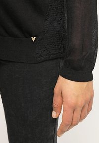 Svart stickad tröja med spetsdetalj på sidan, texturerad tyg och en liten guldfärgad detalj vid fållen.