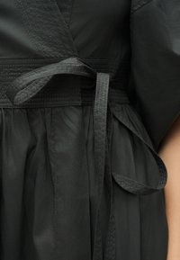 Εστιασμένη εικόνα μαύρου υφασμάτινου φορέματος με υφασμένο ζωνάρι στη μέση και λεπτομέρεια ζώνης δεμένης πάνω από το συγκεντρωμένο τμήμα της φούστας.