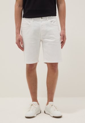 Man draagt witte denim shorts tot op de knie, witte sneakers en een zwart shirt, staand tegen een effen lichte achtergrond.