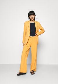 Blazer jaune et pantalons évasés, associés à un top bleu marine et des sandales noires. Texture lisse avec un design ajusté et des lignes épurées.