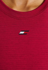Red athletic shirt with a textured surface. Features a small, decorative logo in blue, white, and red located on the chest.