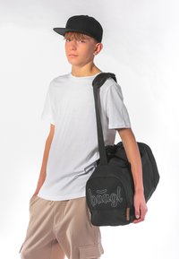 Černá taška přes rameno s zakřiveným tvarem, látkový materiál a šedé logo. Model má na sobě bílé tričko s krátkým rukávem a béžové šortky. Černá kšiltovka.