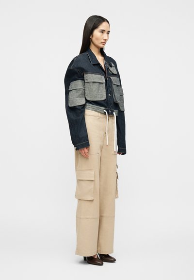 Γυναίκα που στέκεται πλάγια φορώντας cropped σκούρο τζιν μπουφάν με μεγάλες γκρι τσέπες και μπεζ φαρδιά παντελόνια cargo με τσέπες.