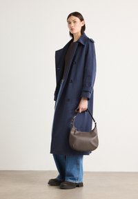 Marineblå trenchcoat, dobbeltradet design, brede slag. Lys brun skinnveske med en buet form. Lyseblå jeans og mørke sko.