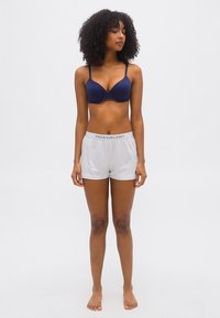 Γυναίκα με σγουρά μαλλιά, όρθια και ξυπόλυτη, φοράει μπλε σουτιέν και λευκά σορτς Polo Ralph Lauren μπροστά σε απλό φόντο.