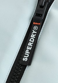 Gros plan sur une fermeture éclair noire sur un tissu clair avec une tirette étiquetée "SUPERDRY®" et un détail de couture orange.