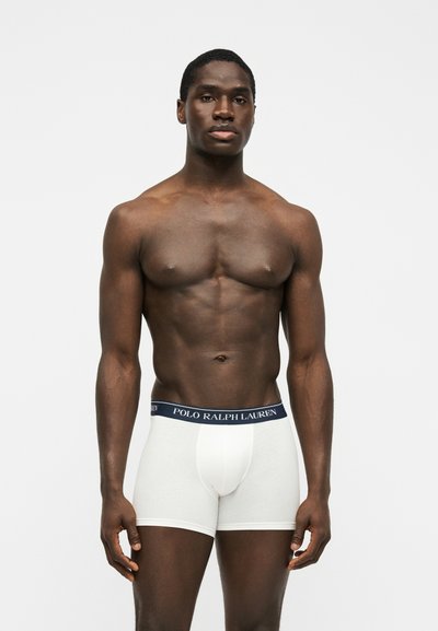 Homme musclé à la peau foncée debout devant un fond blanc, portant un boxer blanc Polo Ralph Lauren, faisant face à l'avant avec les bras détendus.