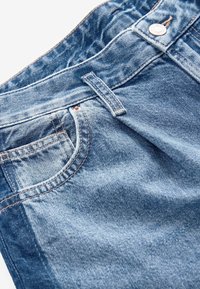 Gros plan sur un jean en denim bleu montrant la poche avant, la passants de ceinture et le bouton métallique avec les détails des coutures visibles.