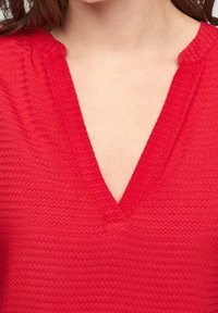 Rød top med korte ærmer og V-udskæring, der har et tekstureret zigzag-mønster og en let afslappet pasform.