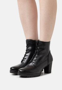 Bottes noires en cuir à la cheville avec un laçage à l'avant, une fermeture éclair sur le côté, un talon épais et une texture lisse. Conviennent pour un usage décontracté ou formel.